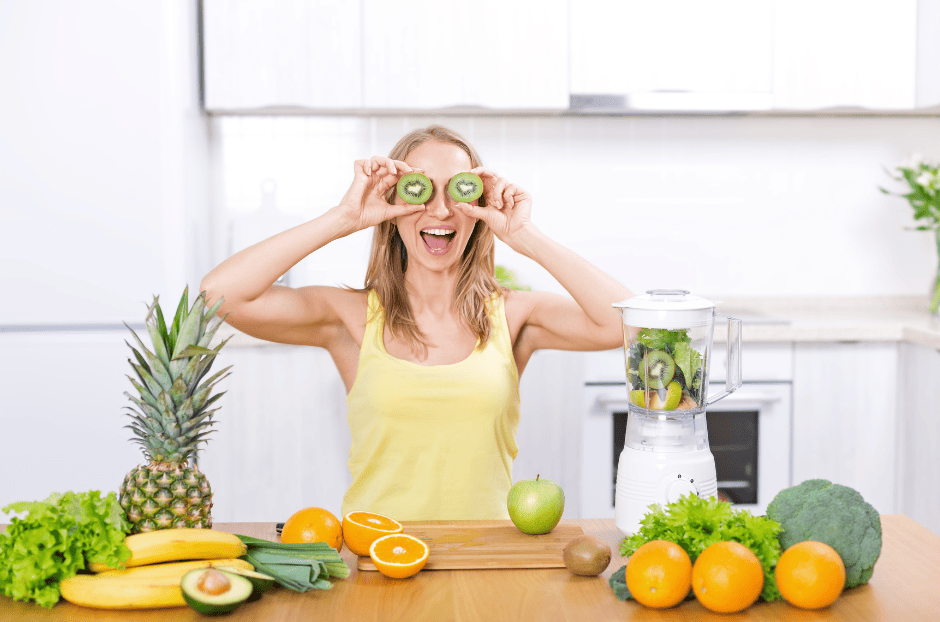 photo d'une femme blanche et blonde cheveu mi-long, elle tient 2 rondelles de kiwis devant ses yeux Sur le plan de travail devant elle, posé à gauche un ananas, des bananes, de la salade, des feuilles de poireaux, une orange entière et deux demis oranges, un demi avocat, à droite, une pomme un kiwi, une salade verte, 3 oranges et un brocoli, un robot avec des fruits et légumes dedans