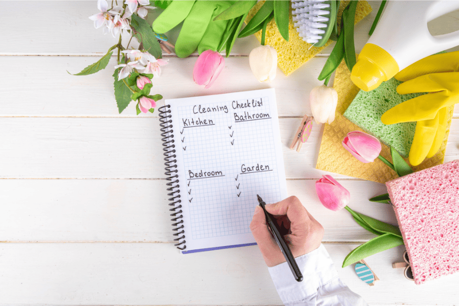 photo d'un carnet à petits carreaux et spirales sur lequel une main écrit avec un stylo noir une check list pour faire le nettoyage, il y a 4 parties une pour la cuisine, une pour la salle de bain, une pour la chambre, une pour le jardin sur le côté droit de l'image on voit une éponge rose et une autre verte posée sur un carré plat de nettoyage jaune, un gant jaune et le haut d'une bouteille blanche avec un bouchon jaune, une brosse verte à poil blanc, une éponge jaune et un gant verte. Des tulipes sont entreposées entre les outils ménagers