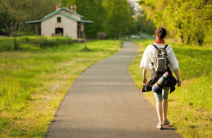 photo d'une femme marchant sur un chemin central, la femme est de dos, elle marche du côté droit et elle porte un sac à dos Au fond du chemin, il y a une maison sur le côté gauche. Le toit est vert et elle est bordé d'arbres.