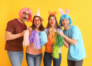 photo de 4 personnes déguisées sur fond jaune en partant de la gauche : homme avec perruque carré rose, lunette ronde noire et fausse moustache, tee-shirt couleur rouille et jean bleu, il tient un pompon rose pâle, femme avec un chapeau pointu à rayures horizontales et grosses pair de lunettes en forme de coeur, cheveu brun mi long, tee-shirt à rayures horizontales oranges et jean bleu, elle tient 2 pompon un violet et un bleu, femme châtain clair cheveu mi long avec un serre-tête papillon, un nez rouge de clown, les yeux qui louchent, un tee-shirt bleu clair, un jean slim bleu, elle tient un pompon orange et un jaune, homme avec perruque carré bleue et 2 oreilles de lapin rose, tee-shirt bleu et jean gris, il tient un pompon vert clair et un vert