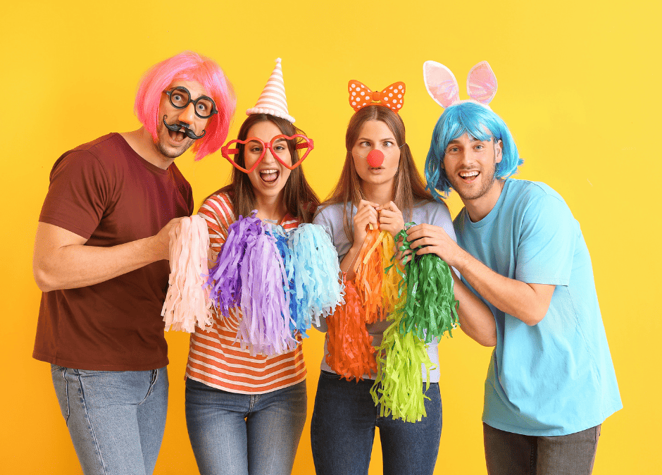 photo de 4 personnes déguisées sur fond jaune en partant de la gauche : homme avec perruque carré rose, lunette ronde noire et fausse moustache, tee-shirt couleur rouille et jean bleu, il tient un pompon rose pâle, femme avec un chapeau pointu à rayures horizontales et grosses pair de lunettes en forme de coeur, cheveu brun mi long, tee-shirt à rayures horizontales oranges et jean bleu, elle tient 2 pompon un violet et un bleu, femme châtain clair cheveu mi long avec un serre-tête papillon, un nez rouge de clown, les yeux qui louchent, un tee-shirt bleu clair, un jean slim bleu, elle tient un pompon orange et un jaune, homme avec perruque carré bleue et 2 oreilles de lapin rose, tee-shirt bleu et jean gris, il tient un pompon vert clair et un vert