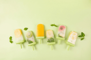 Images de 6 glaces bâton sur fond vert clair. De gauche à droite, glace avec une rondelle de citron glacé en haut, seconde glace avec une rondelle de kiwis glacée, 3ème glace toute orange, 4ème glace avec un citron glacé et des feuilles vertes, 5ème glace avec un morceau de pastèque glacé et dernière glace avec une rondelle de citron glacée