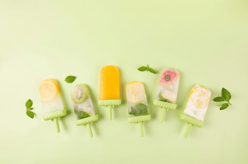 Images de 6 glaces bâton sur fond vert clair. De gauche à droite, glace avec une rondelle de citron glacé en haut, seconde glace avec une rondelle de kiwis glacée, 3ème glace toute orange, 4ème glace avec un citron glacé et des feuilles vertes, 5ème glace avec un morceau de pastèque glacé et dernière glace avec une rondelle de citron glacée