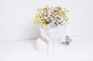 Photo d'un vase blanc en céramique de la forme d'un visage féminin et dont le haut de la tête contient un bouquet de fleurs colorés. Un papillon est posé à droite du vase.