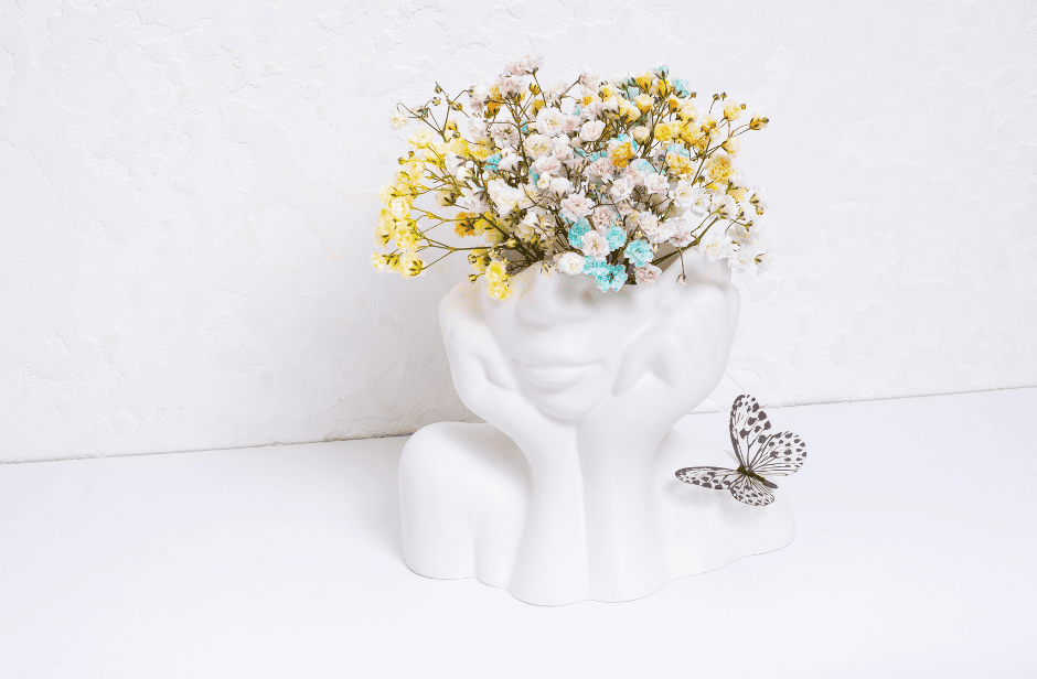 Photo d'un vase blanc en céramique de la forme d'un visage féminin et dont le haut de la tête contient un bouquet de fleurs colorés. Un papillon est posé à droite du vase.
