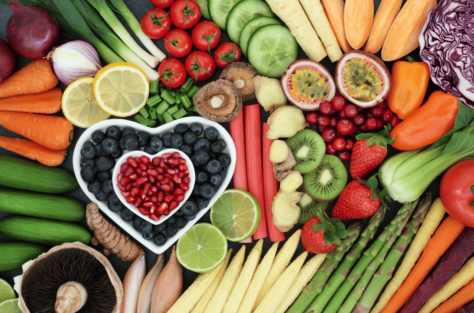 photo de fruits et légumes de l'été avec sur la gauche un plat en forme de coeur de myrtilles, au-dessus un plus petit plat en forme de cœur contenant des dattes. On voit des rondelles de citron, des asperges, des carottes, des tomates, un comcombre tranché, des poireaux, des fraises, un kiwi coupé en 2...