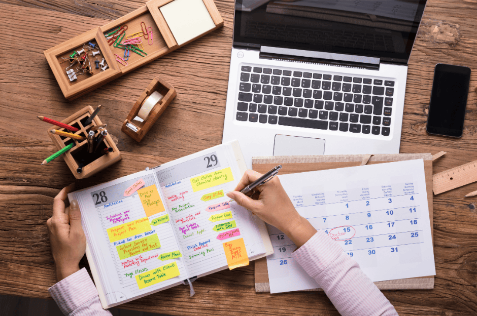 Photo d'un bureau en bois avec un ordinateur portable ouvert, un planning et un agenda où on voit une main de femme écrire.