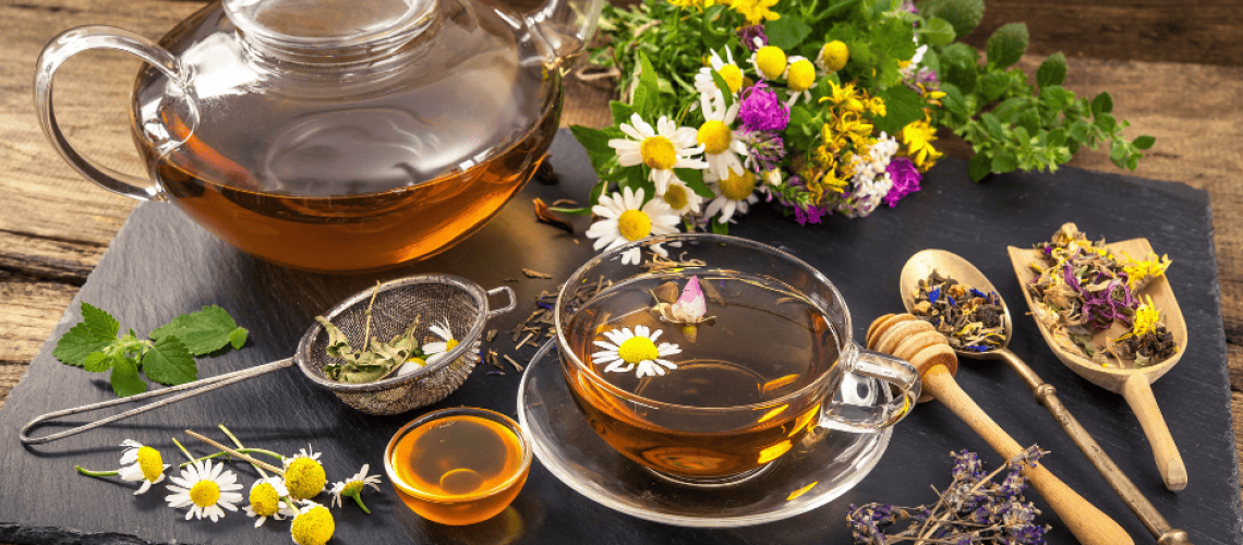 sophrologie-naturopathie-tisane photo d'un plan de travail en bois où est posé sur une ardoise une théière en verre remplie d'un liquide brun entourée de plantes et d'ustensiles (passoire à thé, cuillère à miel, cuillère en argent, cuillère en bois), une tasse remplie posée sur une soucoupe