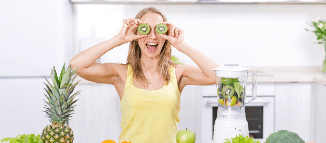 sophrologie-naturopathie-femme-cuisine photo d'une femme blanche et blonde cheveu mi-long, elle tient 2 rondelles de kiwis devant ses yeux Sur le plan de travail devant elle, posé à gauche un ananas, des bananes, de la salade, des feuilles de poireaux, une orange entière et deux demis oranges, un demi avocat, à droite, une pomme un kiwi, une salade verte, 3 oranges et un brocoli, un robot avec des fruits et légumes dedans