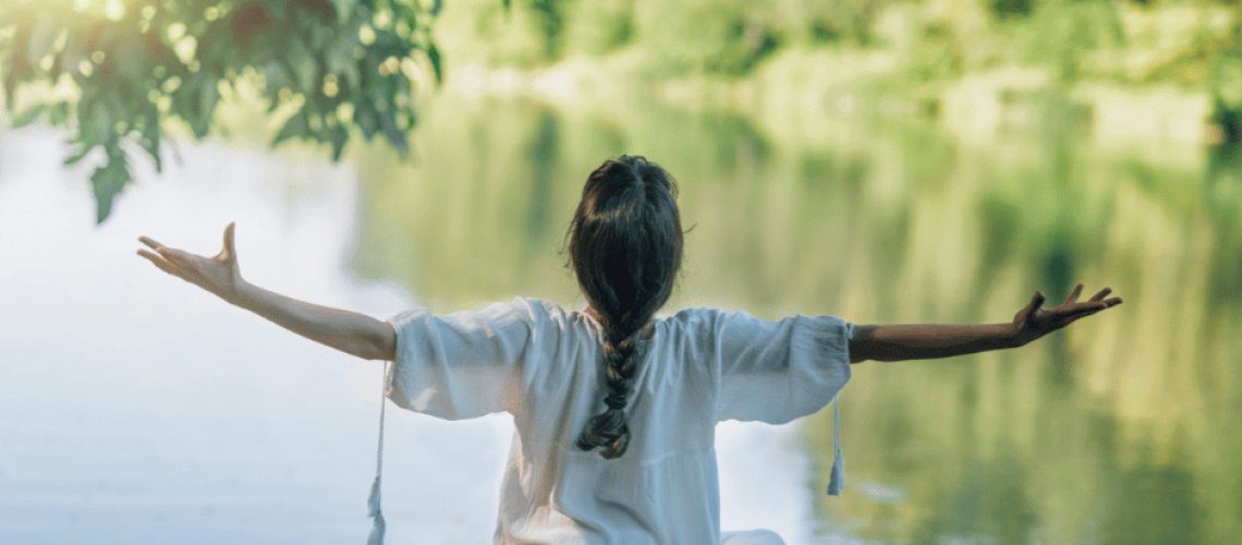 sophrologie-naturopathie-femme-nature photo d'une femme assise de dos au bord de l'eau et qui écartent les bras en haut à droit il y a un branchage perçé par un rayon de soleil