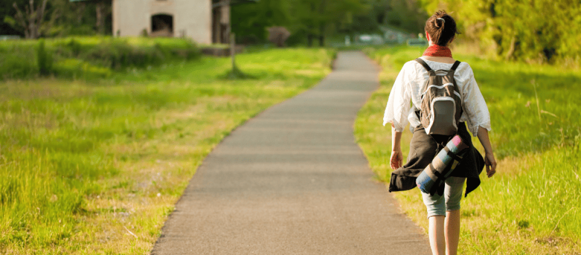 sophrologie-naturopathie-femme-randonnée photo d'une femme marchant sur un chemin central, la femme est de dos, elle marche du côté droit et elle porte un sac à dos Au fond du chemin, il y a une maison sur le côté gauche. Le toit est vert et elle est bordé d'arbres.