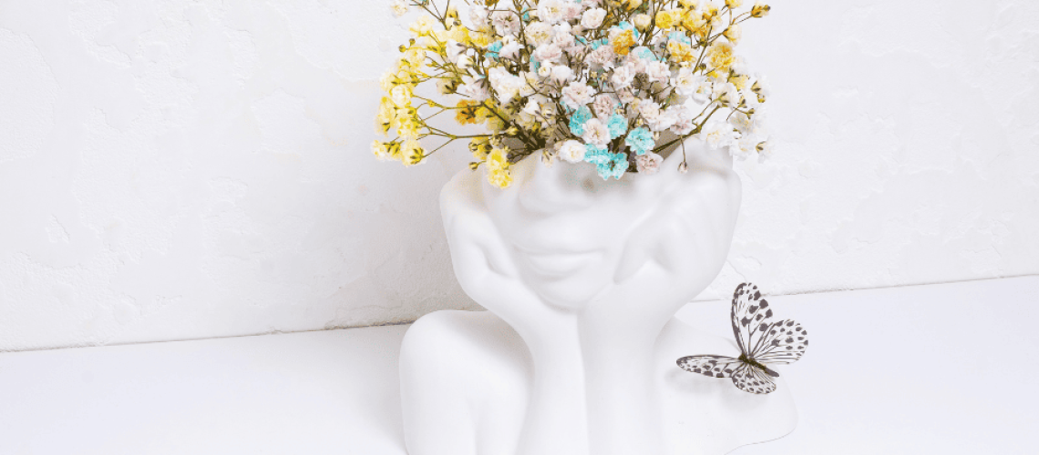 sophrologie-naturopathie-sante-mentale Photo d'un vase blanc en céramique de la forme d'un visage féminin et dont le haut de la tête contient un bouquet de fleurs colorés. Un papillon est posé à droite du vase.