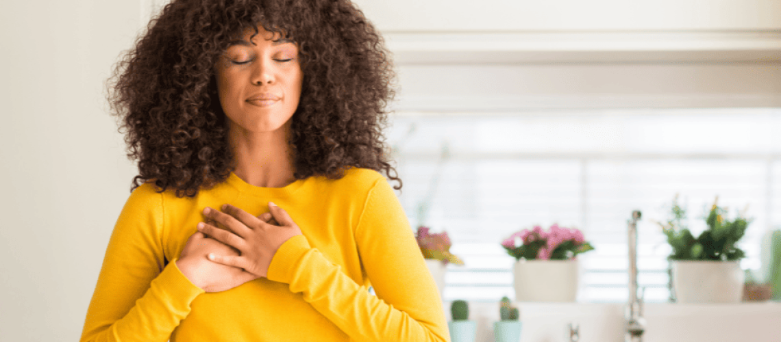sophrologie-naturopathie-femme-gratitude Femme métisse avec une chevelure bouclée, un pull jaune qui tient ses mains posées sur le cœur en signe de remerciement. Elle a les yeux fermés